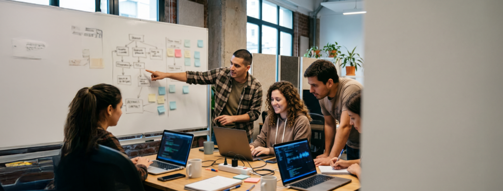 Una persona marca un punto de atención en una pizarra que contiene un gráfico mientras otros compañeros de trabajo desarrollan actividades laborales sobre una mesa con sus computadoras
